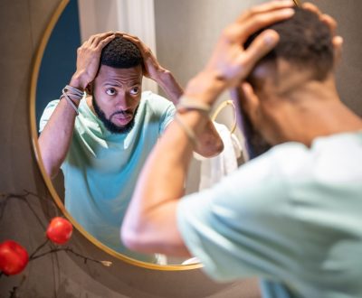 Mens hair loss. Upset young African American man looking in bathroom mirror touching hair, depressed black guy worrying about balding. Male baldness and mental health concept